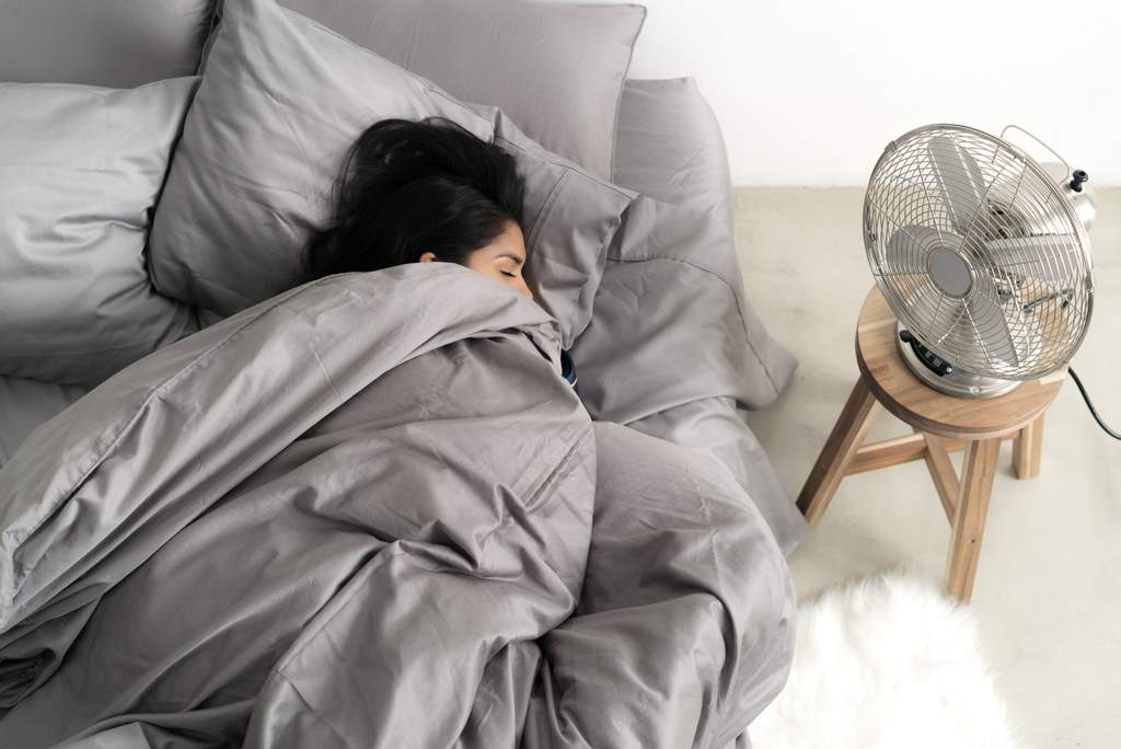 A person sleeping comfortably in bed with a fan running nearby
